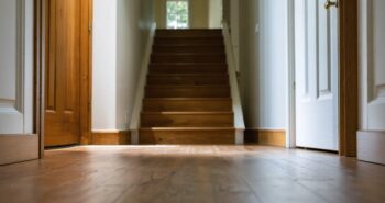 Eye-level view down a modern home hallway with wooden floorboards, highlighting floor seams and a small hairline crack above a white doorframe, lit by soft side daylight with a staircase and living area softly blurred in the background
